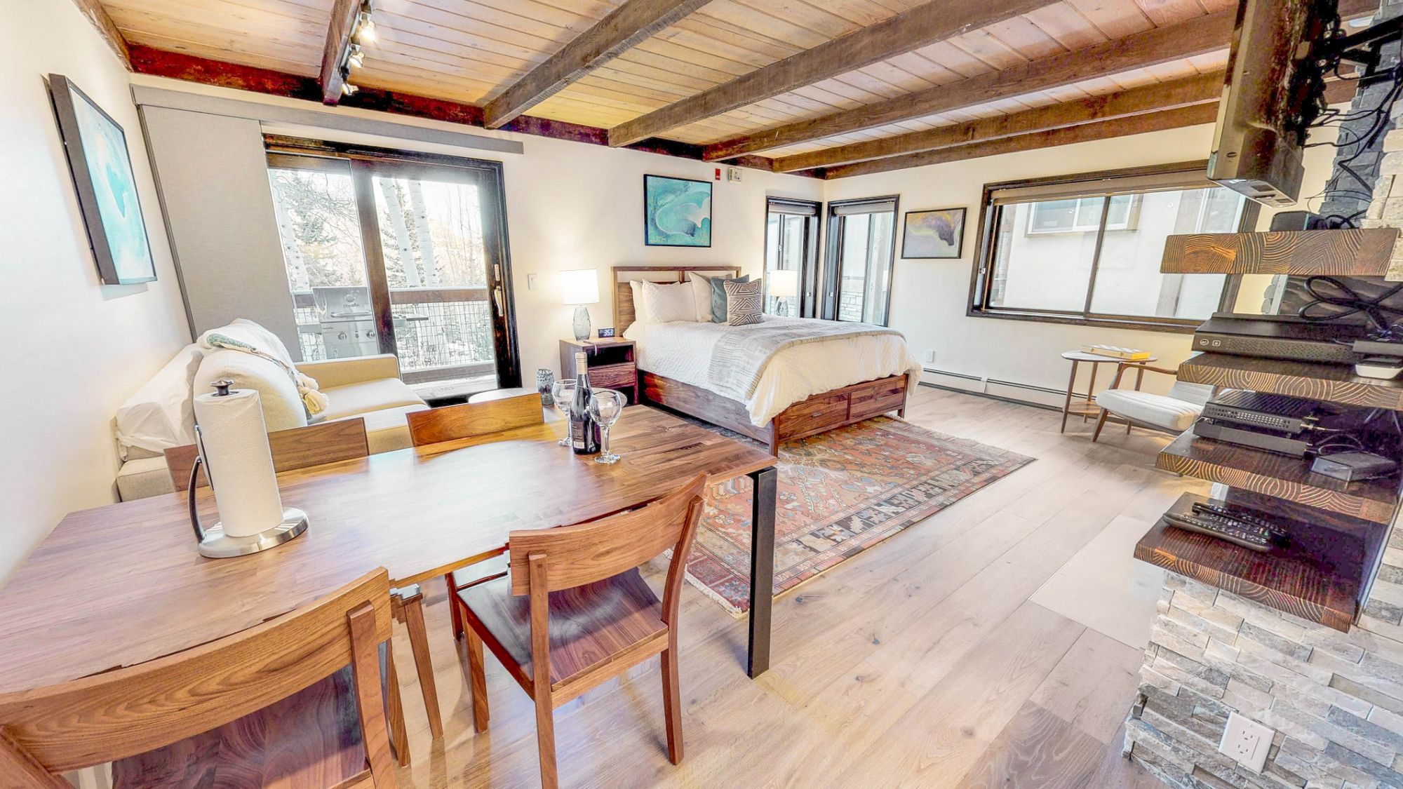 A cozy studio apartment featuring a wooden dining table, a bed, a sofa, and large windows allowing natural light to flood the space.