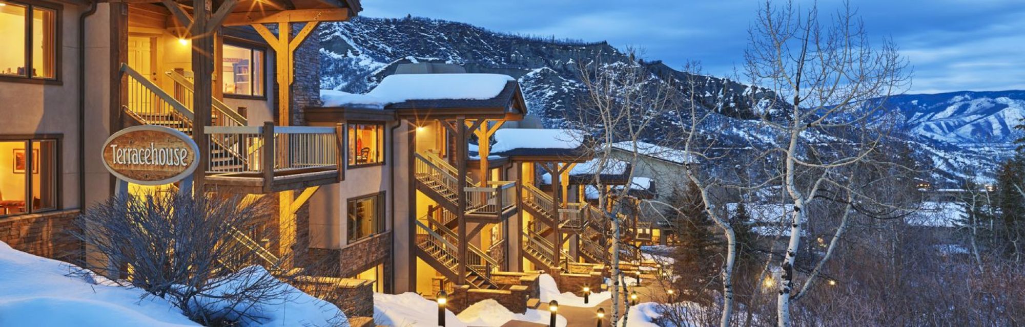 A snowy mountain lodge with lit balconies called Terracehouse, surrounded by barren trees and distant mountain views at dusk.