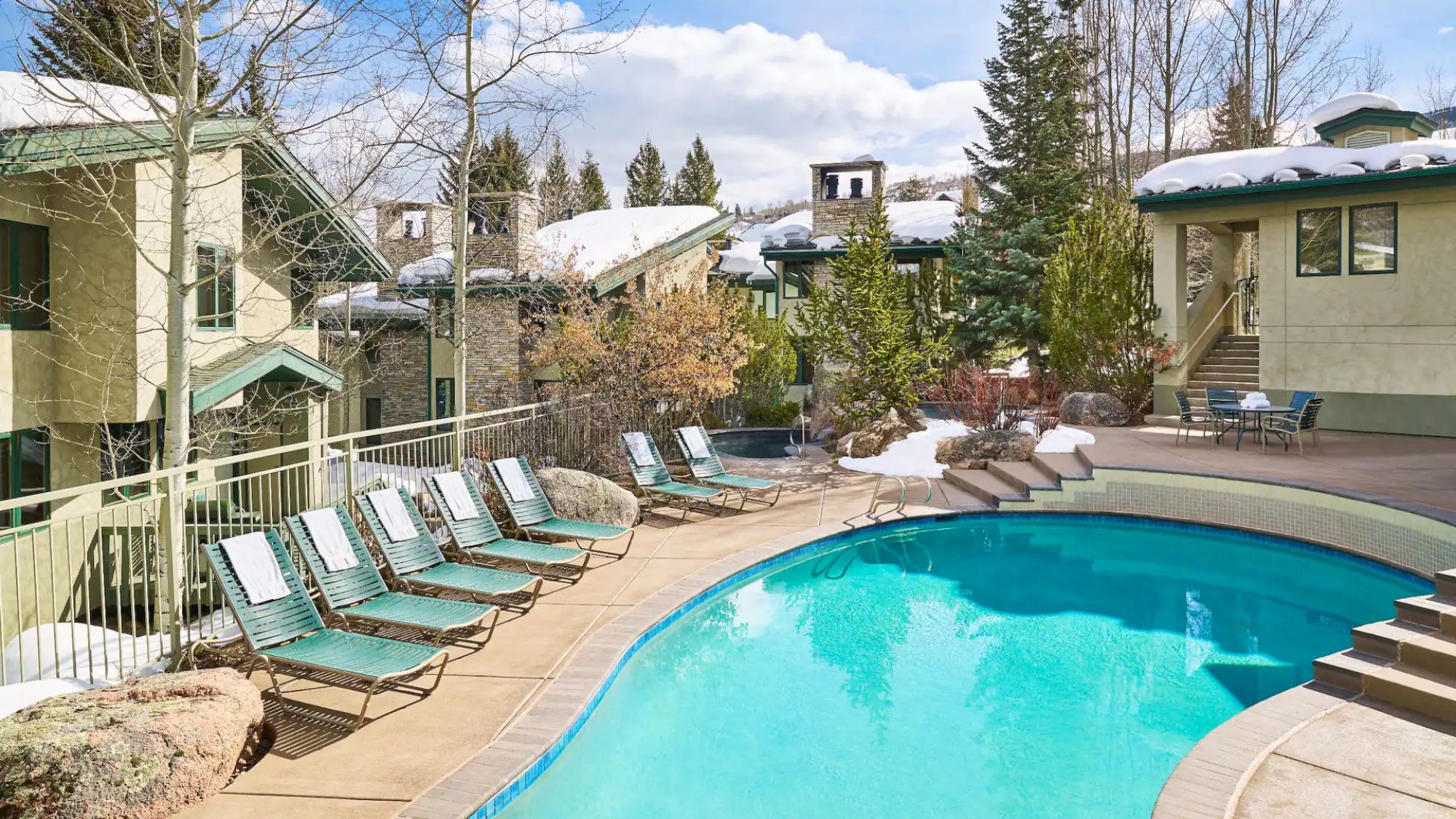 The image features a serene outdoor pool area with several lounge chairs. Snow-capped buildings and trees are visible in the background, under a sunny sky.