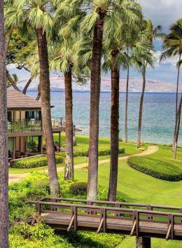 A scenic coastal resort with palm trees, green lawns, and a wooden bridge overlooking the blue ocean.