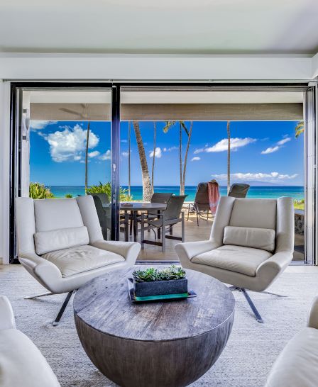 A modern living area with white chairs and a round coffee table, overlooking a patio with ocean and palm trees through large glass doors.