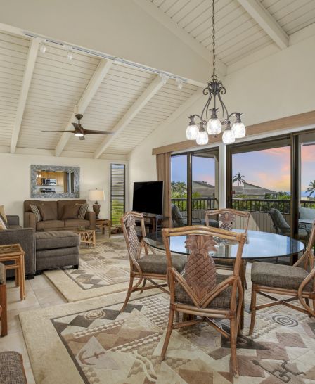 The image shows a spacious room with a dining area, living area, glass doors leading to a balcony, wooden furniture, and rugs on the floor.