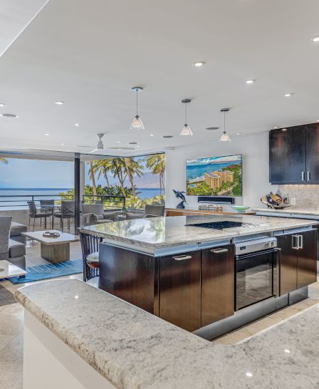 Modern kitchen with granite countertops, stainless steel appliances, and island. Ocean view with palm trees visible through the open sliding doors.