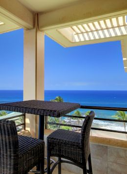 A balcony with a wicker table and chairs overlooks a serene ocean view, with blue sky and palm trees.