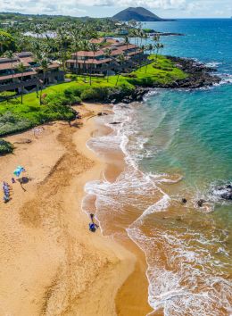 An aerial view of a beach with people under umbrellas, green grassy areas, beachfront buildings, and waves gently lapping at the shore.