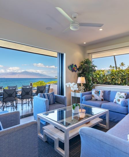 A modern living room with blue sofas and a glass coffee table overlooks a sunny outdoor patio with a stunning ocean view and seating area at a distance.