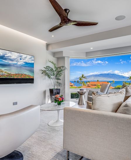 A modern living room with a TV, white furniture, a ceiling fan, and a large window showcasing an ocean view and a clear blue sky dotted with clouds.