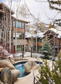 This image shows a winter resort or lodge with wooden buildings, a small hot tub surrounded by rocks, and bare trees under a partly cloudy sky.