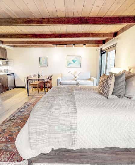 A cozy bedroom with a large bed, fireplace, TV, wooden beams, a rug, artwork, and an open-plan kitchen and dining area in the background.