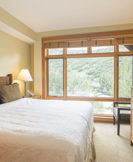 The image shows a cozy bedroom with a large bed, art on the wall, wooden furniture, and a window with a scenic view of green trees and mountains.