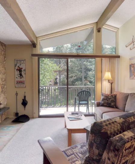 A cozy living room with a stone fireplace, couches, a coffee table, a standing lamp, and wall-mounted decor, opening to a balcony with outdoor seating.