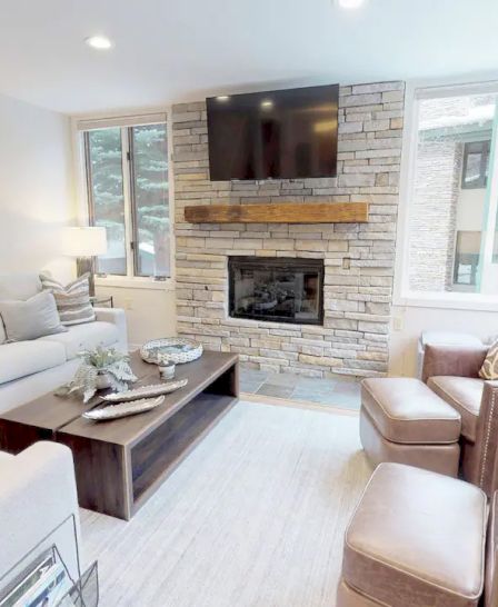 A cozy living room with a stone fireplace, wall-mounted TV, beige sofas, leather chairs, and large windows allowing natural light to fill the space.