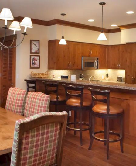 A warm-toned kitchen and dining area with wooden cabinets, plaid-upholstered chairs, a rectangular dining table, and a kitchen island with bar stools.