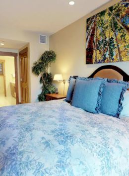 A cozy bedroom features a large bed with blue bedding, wooden furniture, a plant, and a vibrant painting on the wall. There is a door to a bathroom.