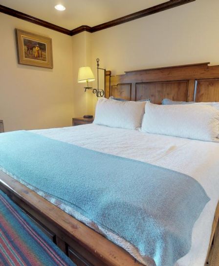 The image shows a cozy bedroom with a wooden bed, blue blanket, two bedside tables with lamps, a window, and a dresser, creating a warm atmosphere.