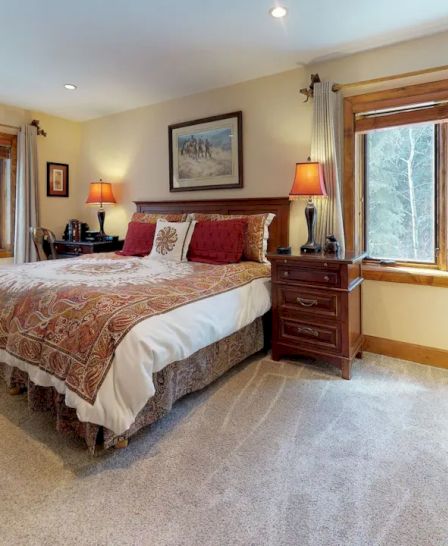 A cozy bedroom with a large bed, two bedside tables with lamps, a wicker chair, and two windows with curtains letting in natural light.