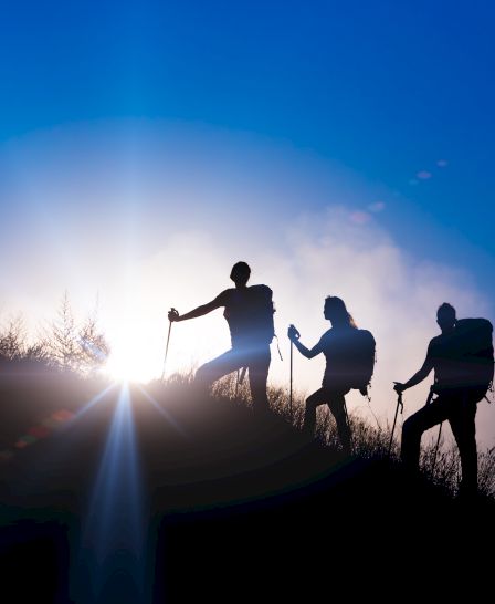 Three hikers with backpacks and hiking poles walking up a hill at sunrise, silhouetted against a vibrant blue sky.