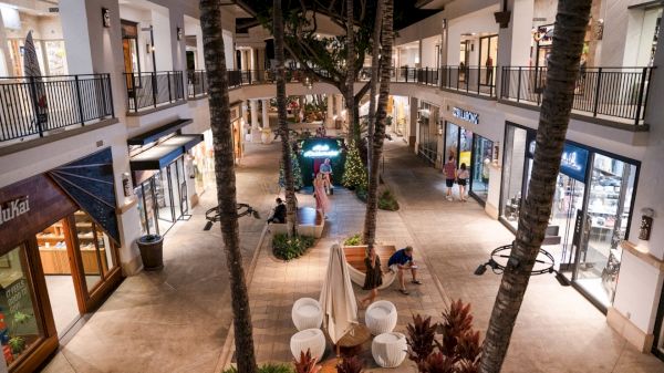 An outdoor shopping center at night with stores on both sides, palm trees, and people walking around.