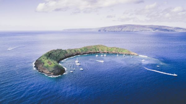 An aerial view of a crescent-shaped island surrounded by blue ocean waters, with boats anchored nearby under a partly cloudy sky.