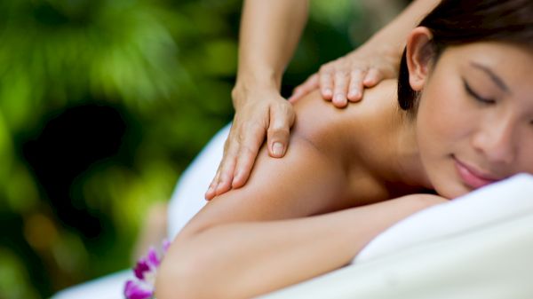 A woman is receiving a relaxing massage with her eyes closed, lying on a massage table outdoors with greenery in the background.