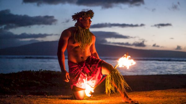 A person dressed in traditional attire is performing a fire dance on a beach at sunset with the ocean and mountains in the background.