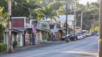 A small-town street with a row of shops, parked cars, and greenery in the background. An American flag hangs on one building, ending the sentence.