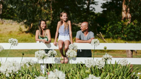 Three people are smiling and sitting on or leaning against a white fence in a garden with white flowers and green foliage in the foreground.