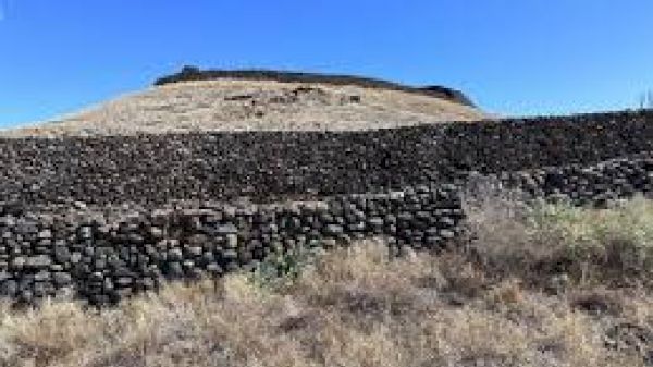 A dry, arid landscape featuring a hill with terraced stone walls under a clear blue sky, and sparse vegetation in the foreground.