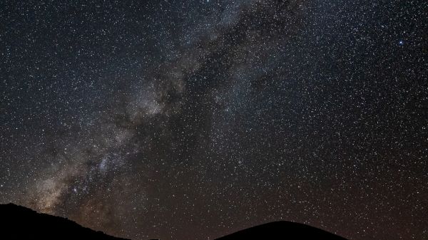 The image shows a night sky filled with stars and a visible section of the Milky Way galaxy, with silhouetted mountains in the foreground.