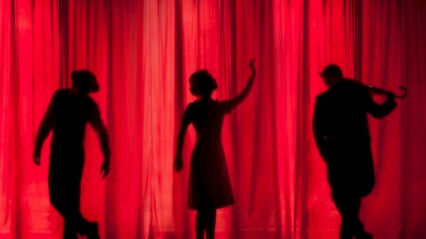 Three silhouetted figures in front of a red curtain appear to be performing or posing dramatically, creating a striking visual against the backdrop.