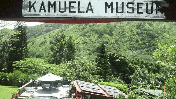 A sign reading "Kamuela Museum" is visible with a lush, green mountainous background and buildings with solar panels and a small pool area.