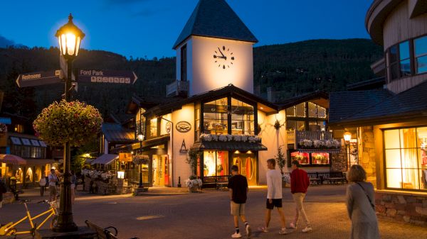 An evening scene of a charming village with a clock tower, street lamps, shops, and people walking on the cobblestone path, surrounded by mountains.