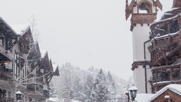 People in winter clothing walk through a snowy, European-style village with chalet-style buildings and a clock tower in the background.