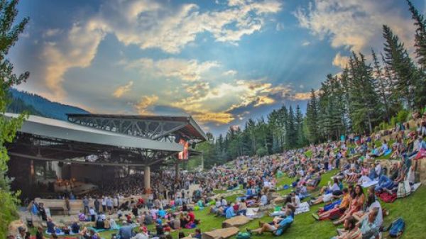 A large crowd is gathered outdoors on a grassy slope with trees, enjoying an event under a clear sky with scattered clouds at sunset.