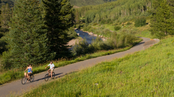 Two people are biking on a paved path surrounded by lush greenery, trees, and a flowing river in the background.