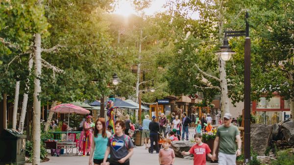 The image shows a lively outdoor area with people walking among trees, shops, and outdoor kiosks on a pleasant day, likely in a park or shopping district.