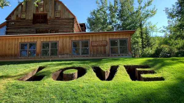 The image shows a wooden building with a grassy lawn in front, where the word "LOVE" is carved into the grass, with trees in the background.