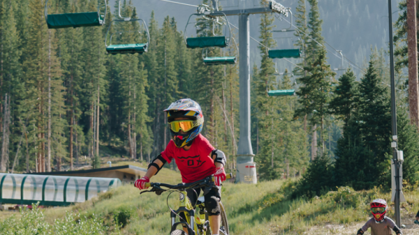 Two children with helmets ride mountain bikes on a wooden path in an outdoor setting with trees, mountains, and a chairlift in the background.