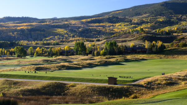 A serene landscape features a lush green golf course set against rolling hills and a clear blue sky, showcasing vibrant autumn foliage in the distance.