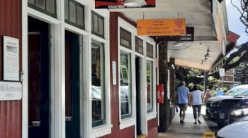 A street view showing a red building with a restaurant, outdoor signage, and a sidewalk with two people walking. Cars and trees are visible.