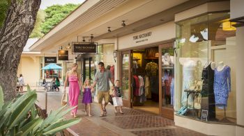 A family is walking through an outdoor shopping area with clothing stores and greenery, carrying shopping bags and enjoying the day.