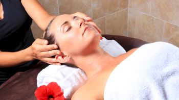 A person is receiving a head massage with eyes closed, lying on a spa bed with a towel and a red flower nearby, looking relaxed.