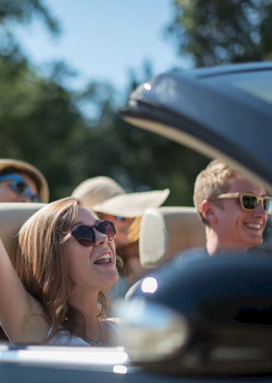 Four people are enjoying a sunny day, riding in a convertible car with the top down, smiling and wearing sunglasses.