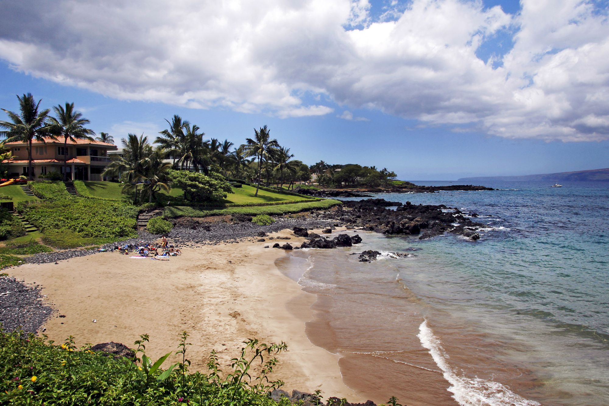 A tropical beach scene with sandy shore, clear blue water, rocky outcrops, and palm trees; homes are visible in the background under a partly cloudy sky.