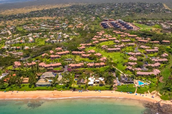 Aerial view of a coastal community with green spaces, residential buildings, and a beach with teal-colored water and some waves along the shore.