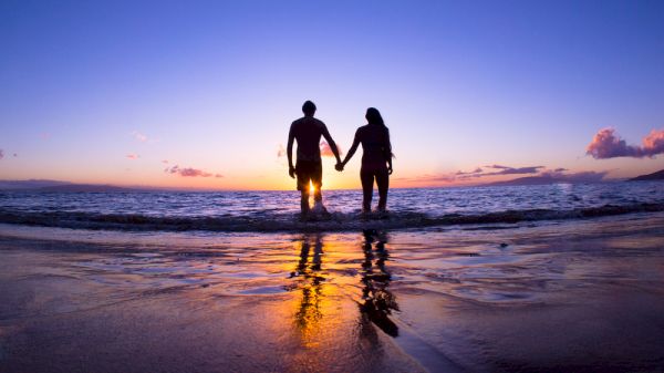 A couple is holding hands while standing in the water during sunset, creating a romantic and serene silhouette against a colorful sky.