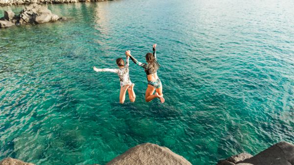 Two people are mid-jump into clear blue water from a rocky shore, holding hands and wearing swimwear, with a sunlit coastline in the background.