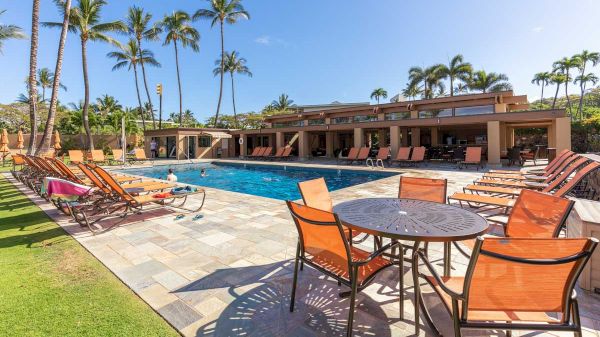 An outdoor pool area with lounge chairs, tables, and palm trees against a clear sky. There is a building in the background, and everything looks sunny.