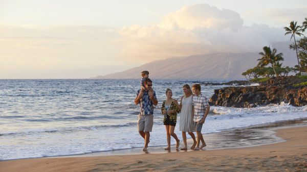 A family is walking on a beach at sunset, with mountains in the background and gentle waves lapping at the shore.