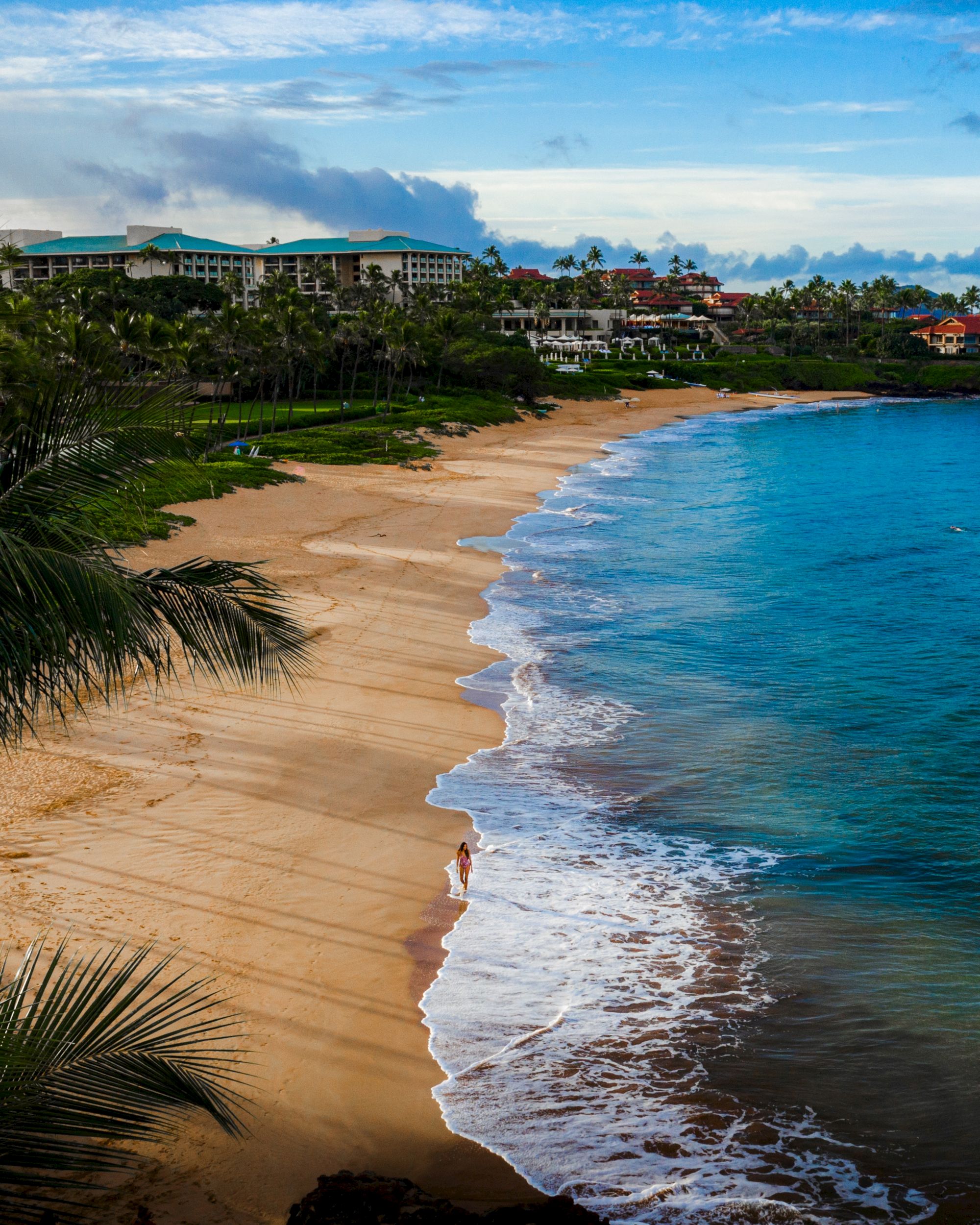 This image shows a tranquil beach with golden sand, clear blue water, and some coastal buildings in the background. The sky is partly cloudy.